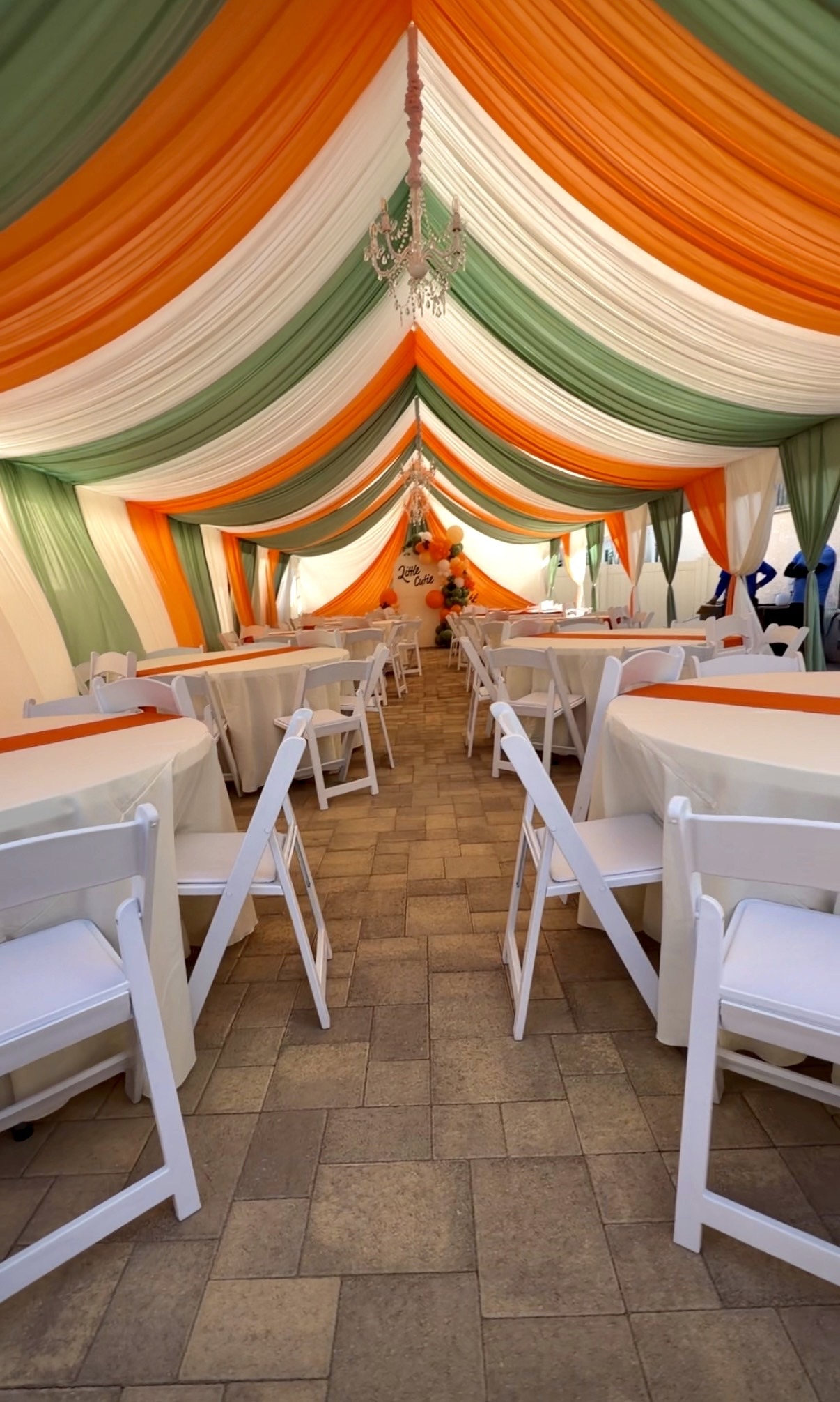 Orange, sage green, and ivory canopy draping with chandeliers, round tables, and white chairs